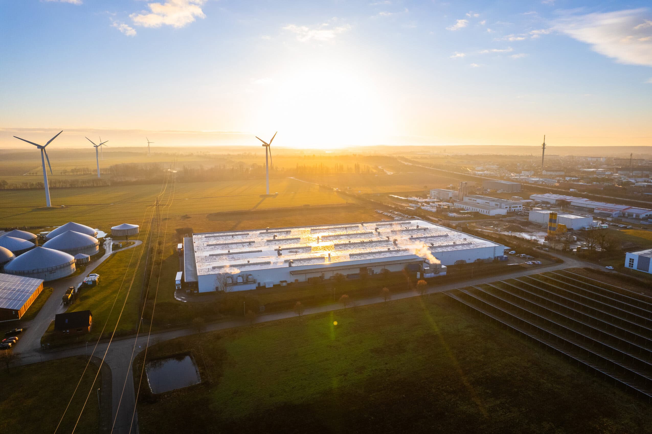 Luftbild Firmengebäude NTN Aerial view of the NTN Antriebstechnik GmbH site in Gardelegen with wind turbines, solar panels, biogas plants, and industrial buildings in a rural setting at sunrise.
