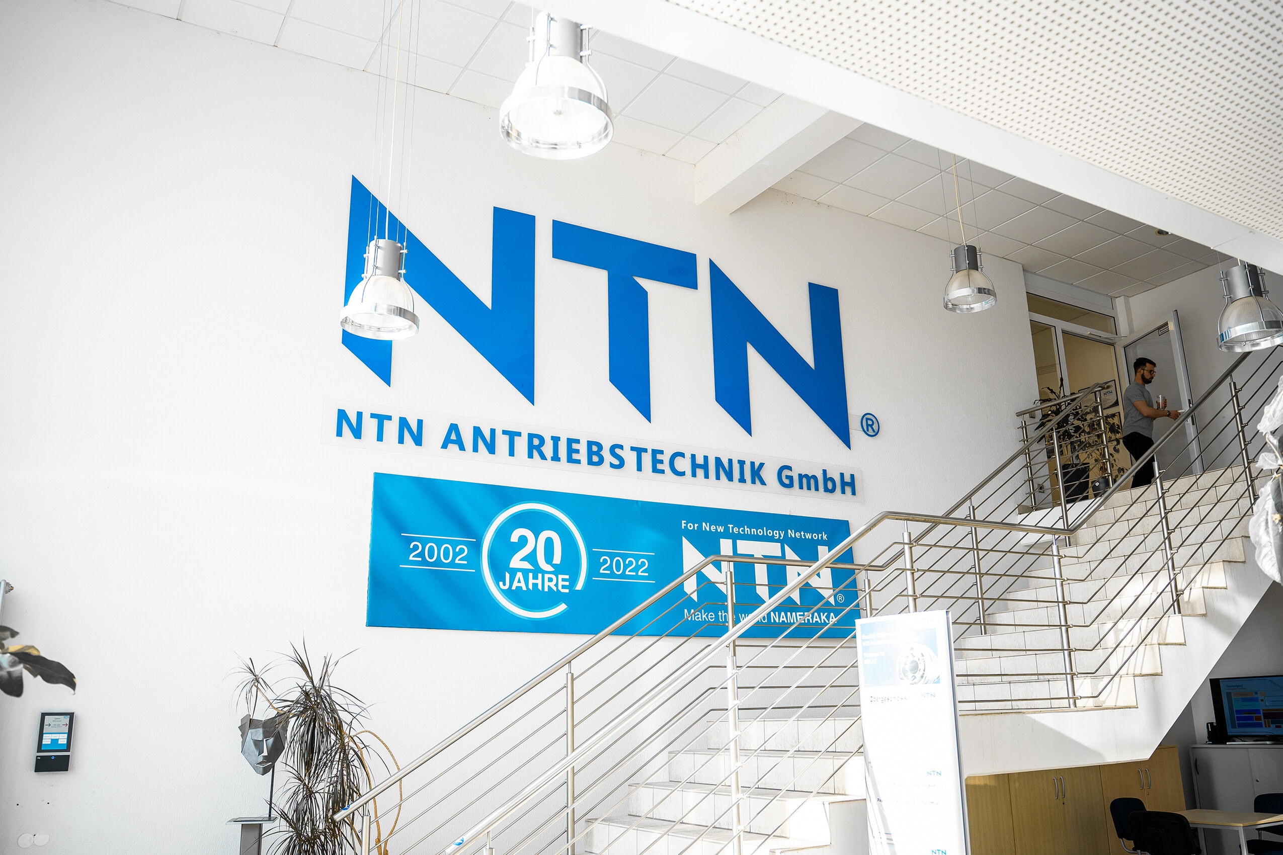 Logo NTN im Treppenaufgang Interior view of the entrance area. Visible are the logo of NTN Antriebstechnik GmbH, the slogan ‘For New Technology Network’, and the anniversary banner ‘20 Years 2002–2022’. Staircase with metal railing, ceiling lighting, and digital info board complete the scene.